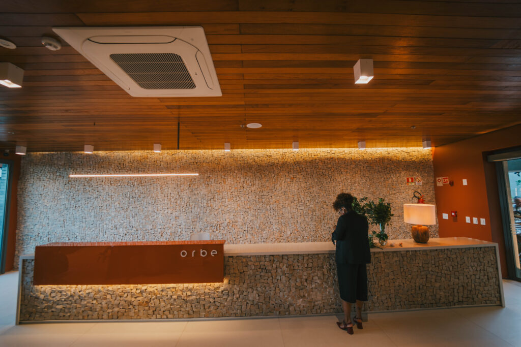 Recepção de design moderno e sofisticado chamada "orbe". O ambiente possui um teto ripado de madeira com iluminação embutida e uma grande parede de fundo feita de pedras claras sobrepostas. O balcão longo tem uma seção frontal na cor terracota com o nome da marca. Uma mulher vestindo roupas pretas está em pé junto ao balcão, próximo a uma luminária de mesa de luz quente.