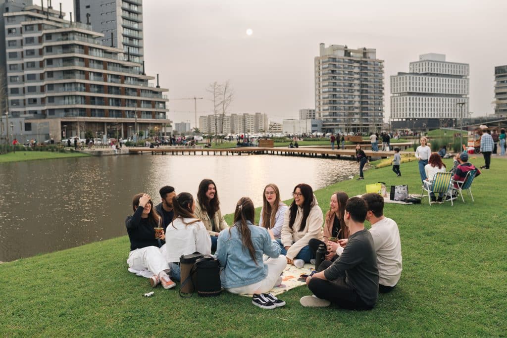 Grupo de jovens sentados em um gramado ao lado de um lago, conversando e compartilhando momentos descontraídos. Ao fundo, edifícios modernos, um píer e outras pessoas aproveitando o espaço ao ar livre, sugerindo um ambiente urbano e comunitário