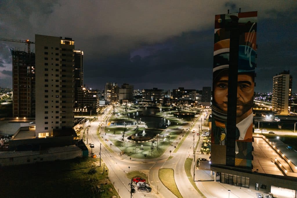 Vista noturna de uma área urbana moderna no Parque Una Pelotas, com ruas iluminadas, um lago central cercado por áreas verdes e edifícios altos, incluindo um prédio com um mural artístico destacado.