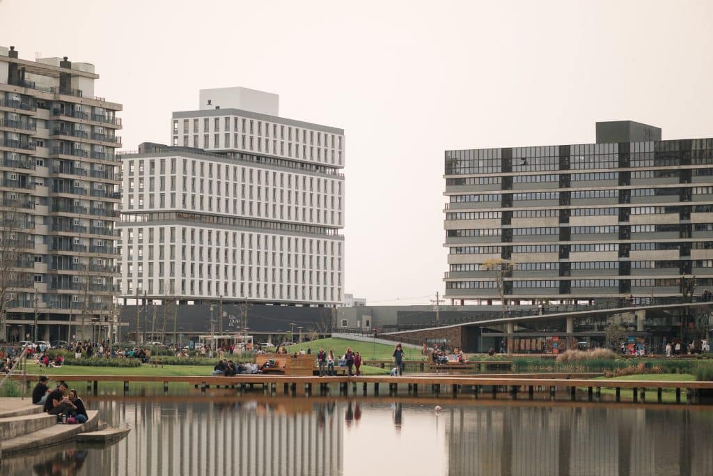 Vista de um parque urbano com um lago refletindo os prédios modernos ao fundo. Pessoas estão sentadas em bancos e gramados, aproveitando o espaço ao ar livre, enquanto a arquitetura contemporânea dos edifícios cria um contraste com o ambiente natural do parque.
