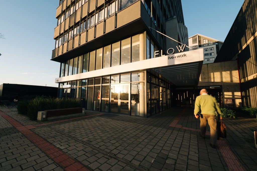 Fachada de um prédio moderno com grandes janelas de vidro refletindo a luz do sol, uma placa com os dizeres 'FLOW live + work' e uma pessoa caminhando em direção à entrada, carregando uma bolsa.