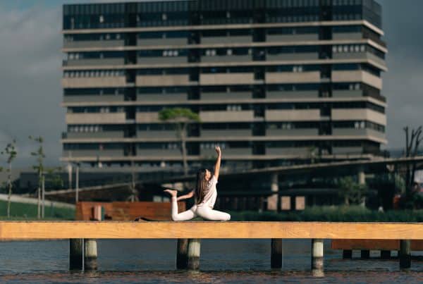 Uma mulher praticando yoga ao ar livre em um píer de madeira com um edifício moderno ao fundo, simbolizando equilíbrio e tranquilidade em meio à urbanidade
