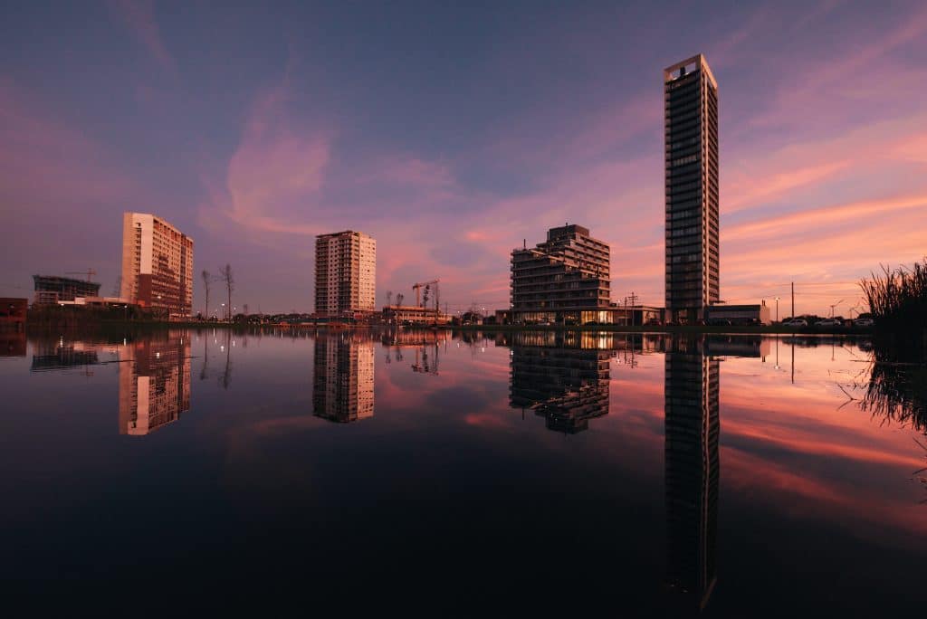 Vista ao entardecer de um complexo moderno de prédios refletidos em um lago calmo, com o céu pintado de tons rosados e roxos, criando uma paisagem urbana serena.