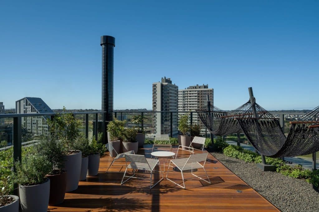 Vista de um terraço com decoração moderna, cadeiras brancas e redes de descanso, rodeado por vasos de plantas e com edifícios residenciais ao fundo em um dia ensolarado.