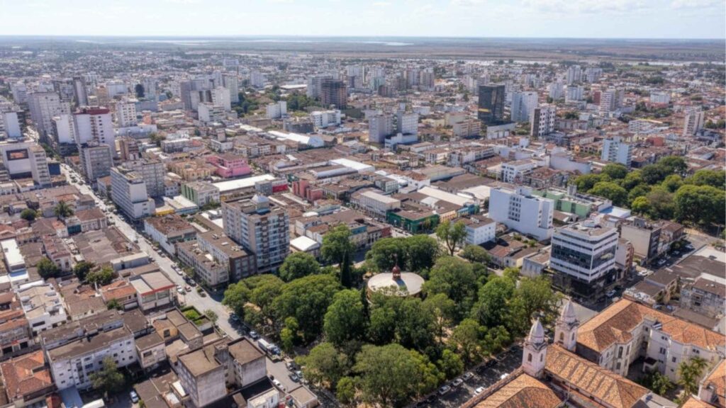 Vista aérea do centro urbano com prédios de diferentes alturas, quadras bem definidas, áreas arborizadas e igreja histórica em destaque, panorama que mostra a diversidade arquitetônica, comercial e residencial dos bairros de Pelotas.