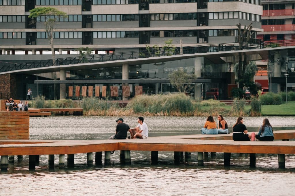 Pessoas sentadas em um deck de madeira sobre um lago no Parque Una, em Uberlândia, durante o fim de tarde. Ao fundo, um prédio moderno com fachada de vidro e vegetação ao redor. O local transmite uma atmosfera tranquila e convidativa para momentos de lazer e convivência.