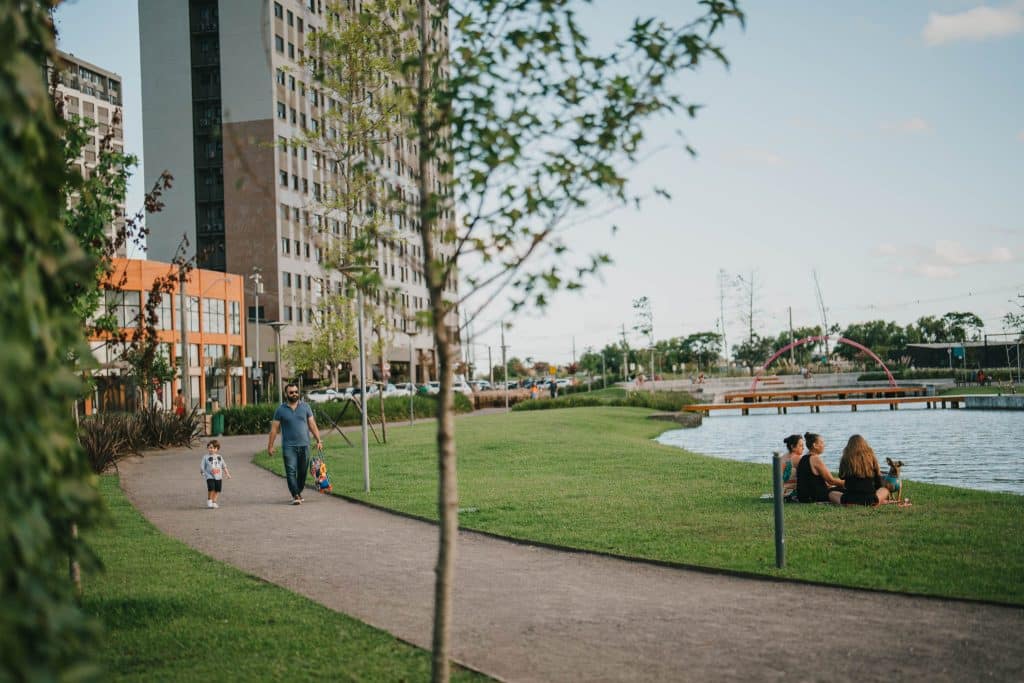 A imagem retrata um parque urbano bem cuidado, com gramado verde, árvores jovens e um caminho de pedestres que contorna um lago. À esquerda, um homem caminha com uma criança pequena, enquanto à direita, três mulheres estão sentadas na grama à beira d’água, acompanhadas de um cachorro, desfrutando de um momento de lazer. Ao fundo, vê-se uma ponte com estrutura em arco vermelho, pessoas passeando e prédios residenciais e comerciais, formando um contraste entre a natureza e a urbanização.