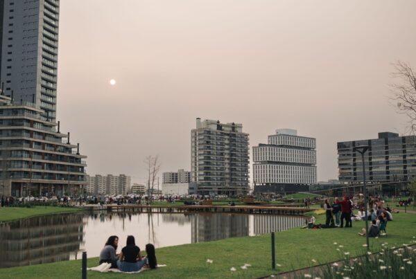 Vista panorâmica de um parque urbano moderno em Pelotas ao entardecer, com um lago artificial no centro e pessoas sentadas no gramado. Ao fundo, destacam-se edifícios residenciais altos e contemporâneos de um bairro planejado.
