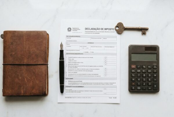 Vista de cima de uma mesa de mármore branco contendo itens de escritório. No centro, há um formulário impresso com o título "DECLARAÇÃO DE IMPOSTO" e uma chave antiga pousada sobre ele. À esquerda do papel, há um caderno com capa de couro marrom rústico e uma caneta-tinteiro preta. À direita, há uma calculadora preta.