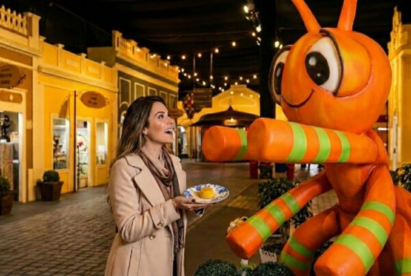 Uma mulher sorridente, vestindo um casaco de inverno bege e cachecol, interage alegremente com a estátua de uma formiga gigante, laranja com listras verdes. Ela segura um pequeno prato com um doce amarelo, parecido com um quindim. O cenário ao fundo é um ambiente fechado decorado como uma vila antiga, com fachadas de casinhas coloridas em amarelo e vermelho e iluminação quente de cordões de luzes no teto.