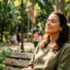 Close-up de uma mulher de olhos fechados e expressão serena, sentada em um banco de madeira em um parque. Ela parece respirar profundamente e aproveitar o sol que filtra entre as árvores ao fundo. O ambiente está desfocado, destacando sua sensação de paz e conexão com a natureza.