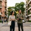 Um casal parado em uma calçada urbana, posicionado entre dois prédios residenciais. No prédio à esquerda, destaca-se uma placa laranja escrito "ALUGA-SE". No prédio à direita, uma placa azul indica "VENDE-SE". O homem segura um pequeno bloco de notas e o casal observa os prédios com uma expressão de dúvida e análise.