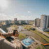 Vista em perspectiva de uma mulher de costas, segurando uma caneca escura em uma sacada de andar alto. Ela observa um bairro planejado e ensolarado, que conta com prédios modernos, uma quadra poliesportiva nas cores azul e laranja, e um lago artificial sinuoso que corta a área verde. O céu está limpo e azul.